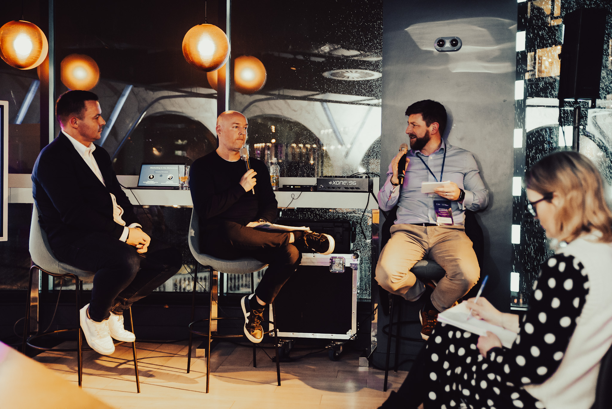 Panelists Greg and Robert sit on stools, with Robert holding a microphone. Host Ewan is also holding a microphone and is asking a question